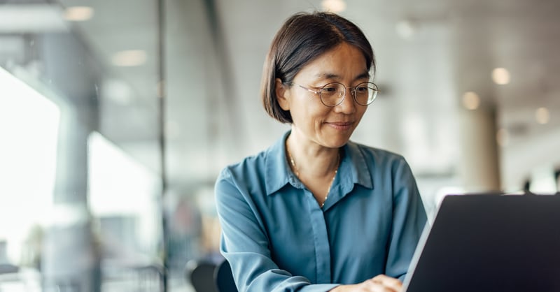 Asian female employee with glasses a blue shirt, sitting at and smiling at her laptop. 