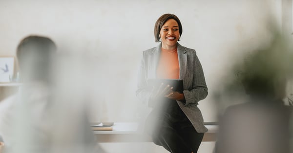 Black woman leaning against a desk, holding a tablet and smiling