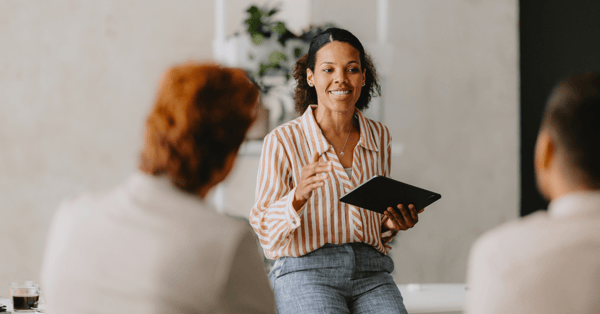 Black business woman leading her team in a discussion, sat on a desk and smiling.