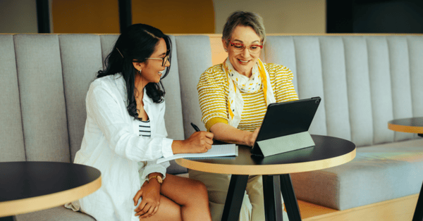 Senior professional woman, wearing a yellow top and scarf, sat at a circular desk, providing a younger woman in a white shirt with coaching support.