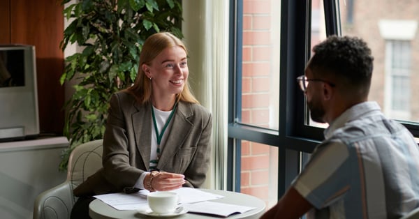 Female manager having a conversation with her male colleague at a small desk, smiling and holding a pen as she takes notes.