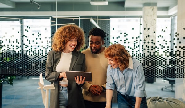 Three employees - two women and one man - standing together and smiling while looking at a tablet. 