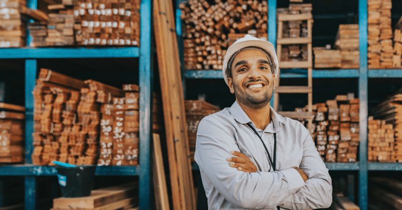 Male lumber supply employee wearing a shirt and hard-hat, stood in a factory setting, crossing his arms and smiling at the camera.