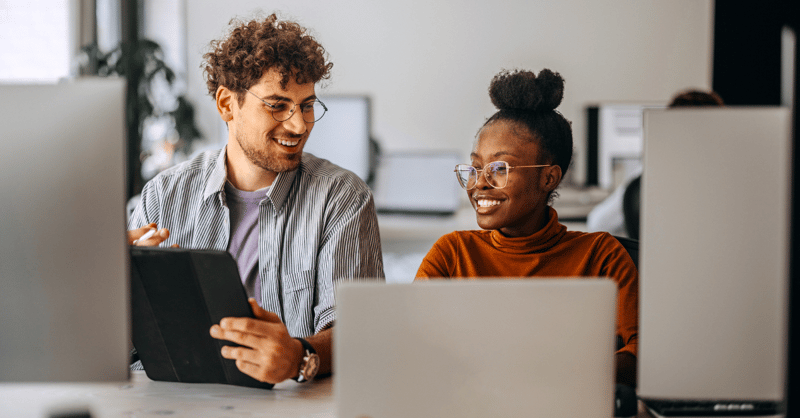 Two employees, one male, one female, sat next to each other in front of computer screens and smiling at each other.