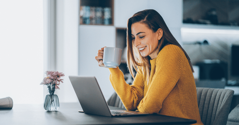 Female employee wearing a yellow jumper and holding a cup of coffee while smiling at her laptop.